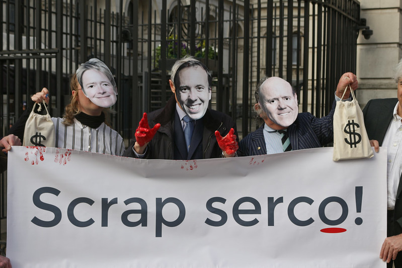 An image of people wearing facemasks of Dido Harding, Matt Hancock and Rupert Soames holding a banner which says 'Scrap Serco'.