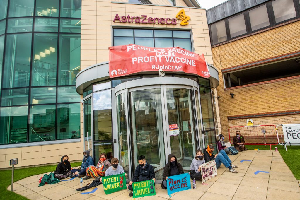 Protesters sitting outside the AstraZeneca office with a banner which says "End vaccine apartheid. We demand a people's vaccine!"