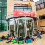 Protesters sitting outside the AstraZeneca office with a banner which says "End vaccine apartheid. We demand a people's vaccine!"