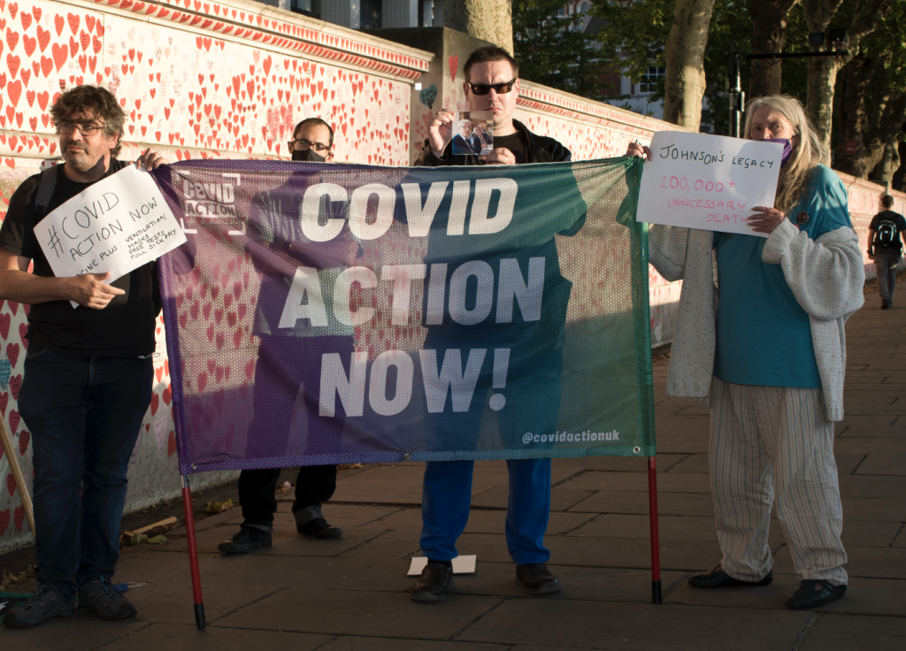 Covid Action activists stand with a banner reading COVID ACTION NOW in front of the memorial wall in central london.