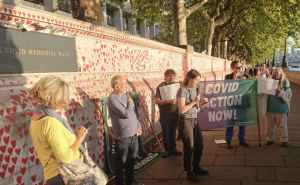 Activists in front of the covid memorial wall listen to a speech holding a covid action now banner