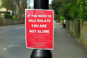 Poster attached to a lamposr. Red background with white writing: If you need to self isolate you are not alone with contact for local mutual aid group and a list of the sort of services they may be able to provide